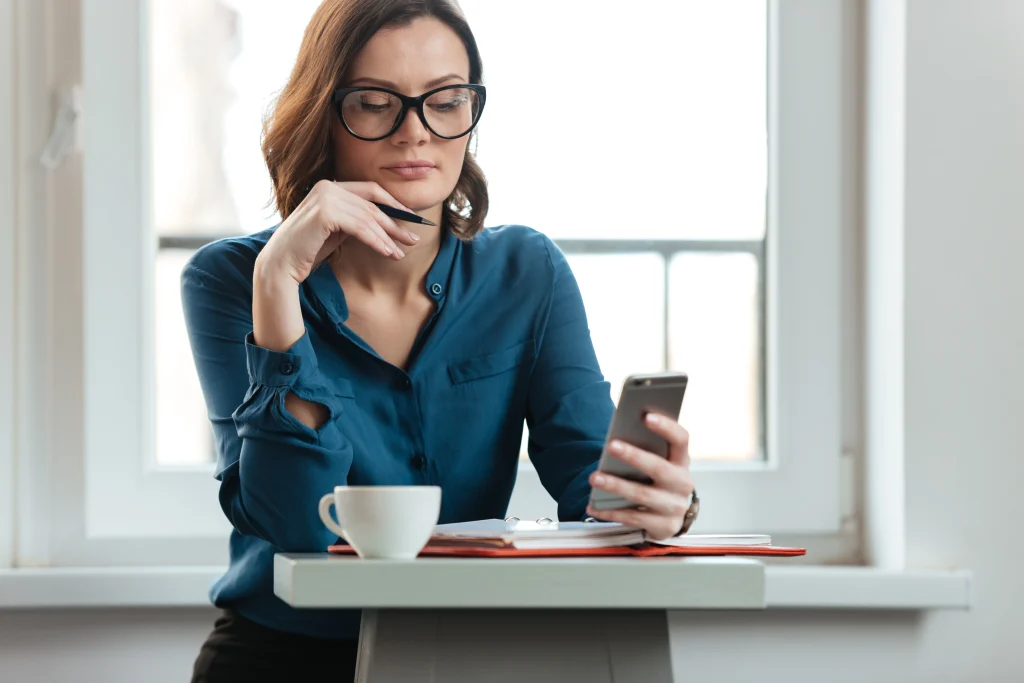Używanie telefonu komórkowego w pracy – kobieta przegląda telefon przy stoliku z notatnikiem