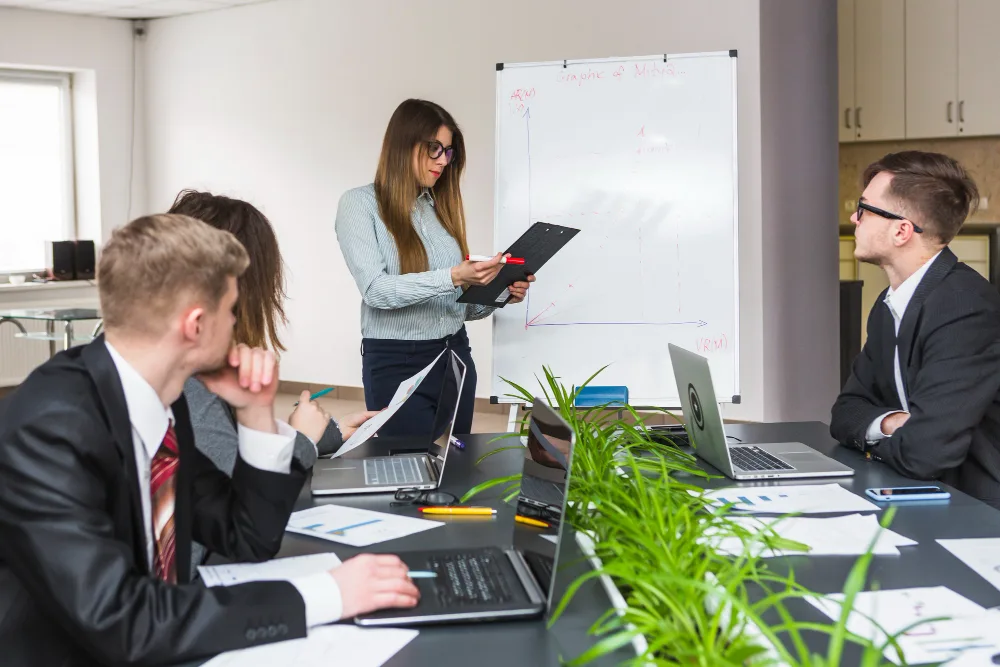 Szkolenia stacjonarne dla księgowych – prowadząca z podkładką przy tablicy, troje uczestników przy stole z laptopami
